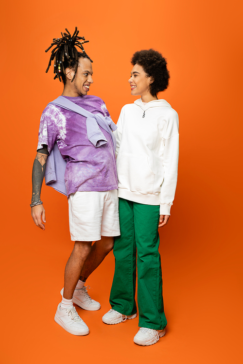 full length of cheerful african american woman and stylish man with dreadlocks looking at each other on orange