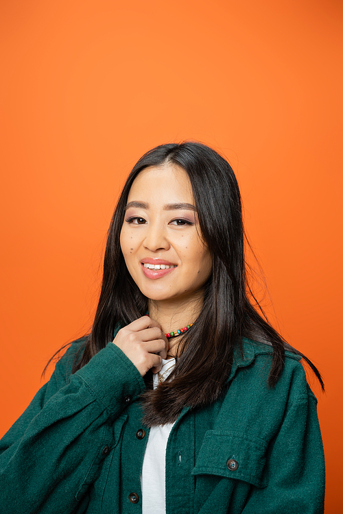 portrait of pretty asian woman in green shirt touching colorful beads and smiling at camera isolated on orange