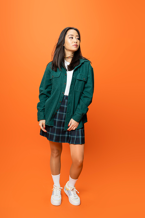 full length of young asian woman in checkered skirt and green shirt looking away on orange background