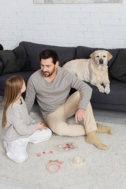 Labrador dog lying on couch near father and daughter sitting on floor near toy crowns and makeup palette