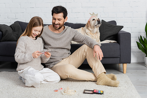 happy father and daughter looking at makeup palette while sitting near labrador dog in living room