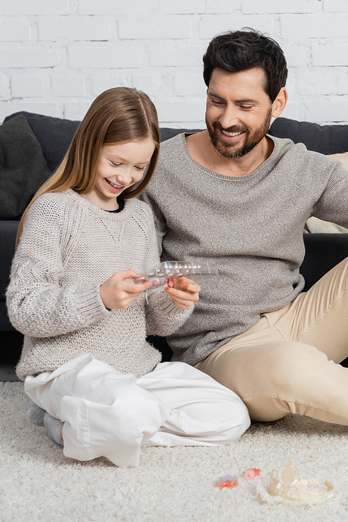 happy father and daughter looking at makeup palette while sitting on carpet near sofa