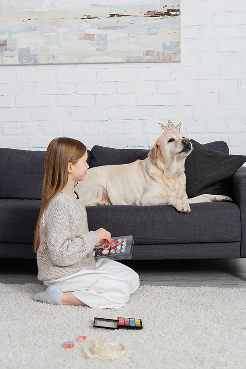 happy preteen girl holding makeup palette and looking at labrador in toy crown sitting on couch