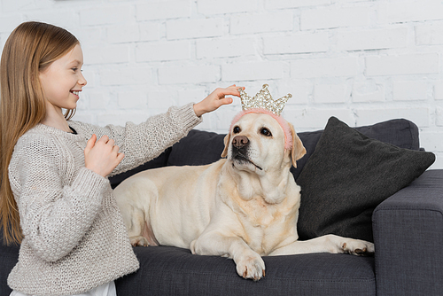 happy preteen girl adjusting toy crown on labrador lying on couch