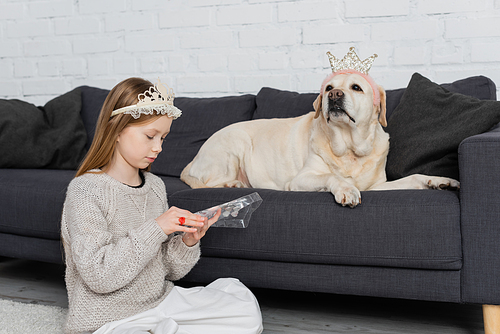 preteen girl in crown looking at makeup palette while sitting near labrador on couch