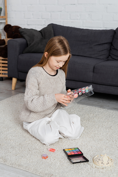 happy preteen girl looking at makeup palette and sitting on carpet near couch