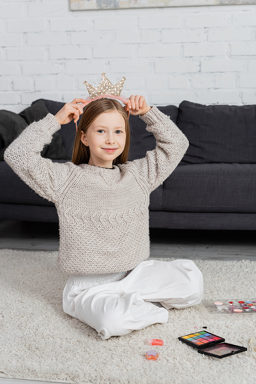 happy preteen girl adjusting toy crown while sitting on carpet near makeup palette