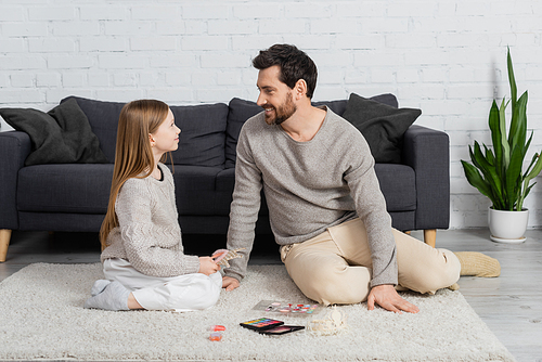 happy father looking at smiling daughter while sitting on carpet near decorative cosmetics