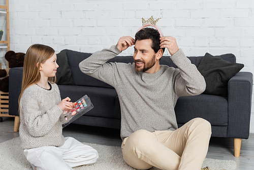 happy man wearing toy crown near cheerful preteen daughter holding makeup palette