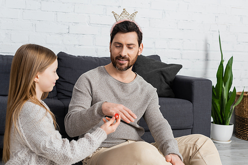 happy girl wearing toy ring on finger of father in crown while playing in living room