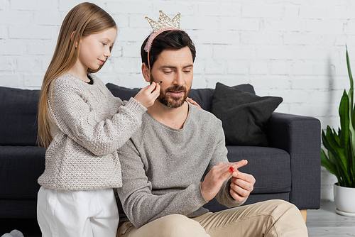 girl holding cosmetic brush while applying makeup on cheek of smiling father in crown