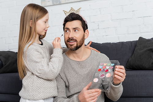 girl holding cosmetic brush while applying makeup on face of smiling father in crown