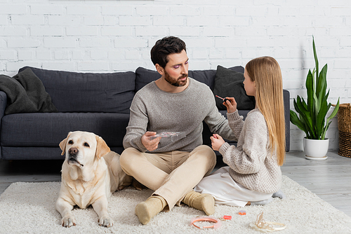 girl holding lip gloss while sitting near father and labrador on carpet