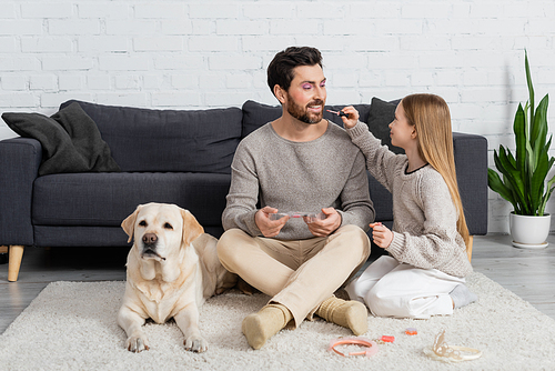 happy girl applying lip gloss on lips of father while sitting with labrador on carpet