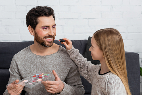 happy girl applying lip gloss on lips of father holding makeup palette