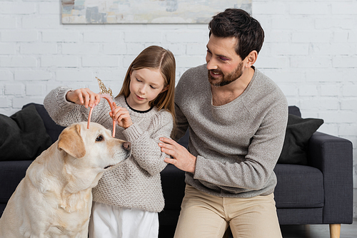 happy girl wearing toy crown on head of labrador near father in living room