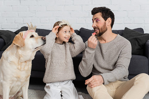 happy girl wearing toy crown on head while sitting near labrador and looking at father applying lip gloss