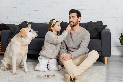 happy girl in toy crown on head sitting near labrador and applying lip gloss on lips of father