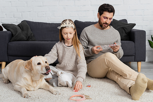 preteen girl in toy crown on head sitting near labrador and father while playing beauty game in living room