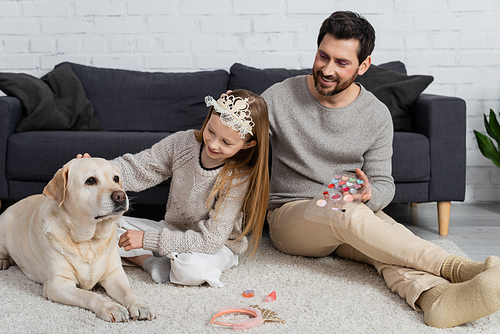 happy girl in toy crown on head cuddling labrador near happy father and makeup products on carpet