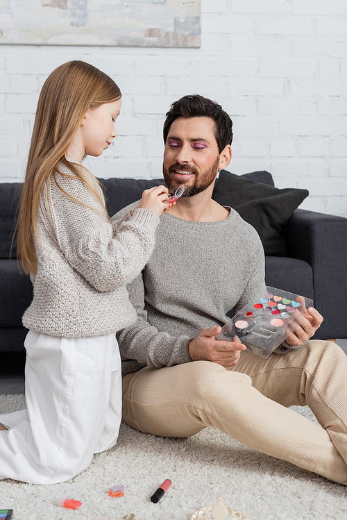 happy father looking at preteen kid holding eye shadow product while playing beauty game