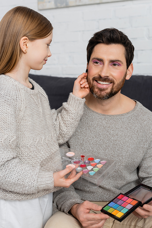 preteen girl holding cosmetic brush while applying beauty product on cheek of bearded father