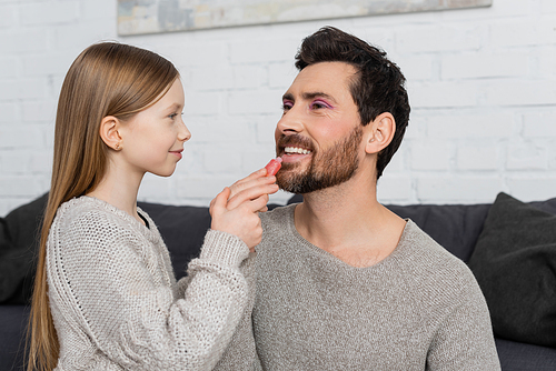 happy preteen girl applying shiny lip gloss on lips of smiling and bearded father