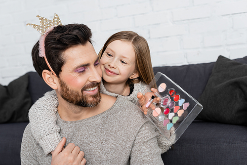 happy kid holding beauty palette and hugging smiling father in toy crown