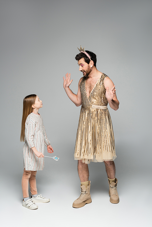 funny father in dress and crown gesturing while looking at daughter with toy wand on grey