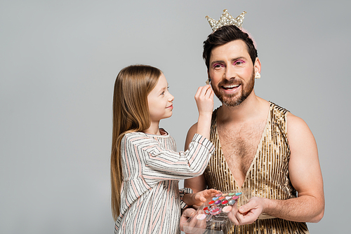 happy kid applying decorative cosmetics on face of bearded father in crown and dress isolated on grey