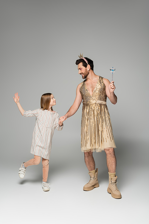 full length of girl in dress holding hands with dad in dress and toy wand on grey