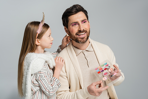 happy girl touching earring of cheerful father with eye shadow palette isolated on grey