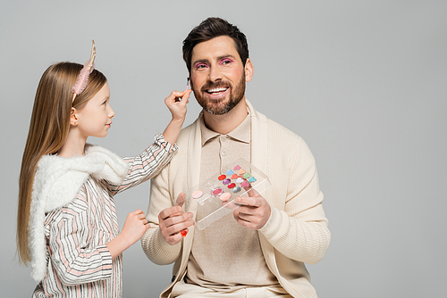 happy girl holding cosmetic brush near cheerful father with eye shadow palette isolated on grey