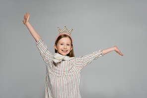 happy girl in dress and crown smiling while standing with outstretched hands isolated on grey