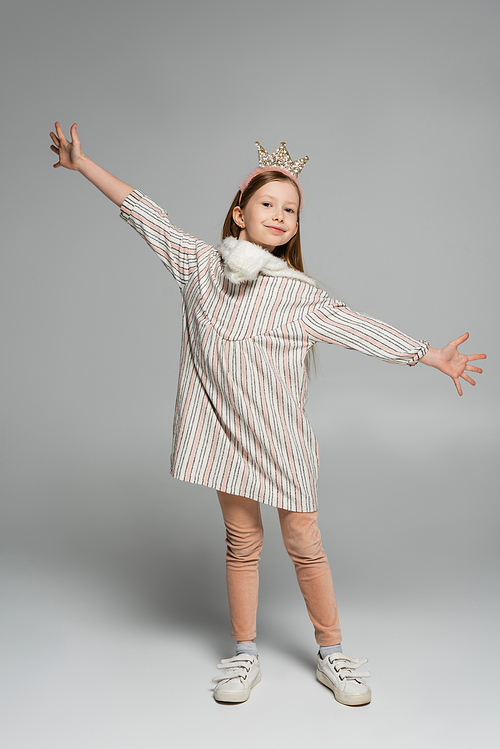 full length of happy girl in dress and crown smiling while standing with outstretched hands on grey