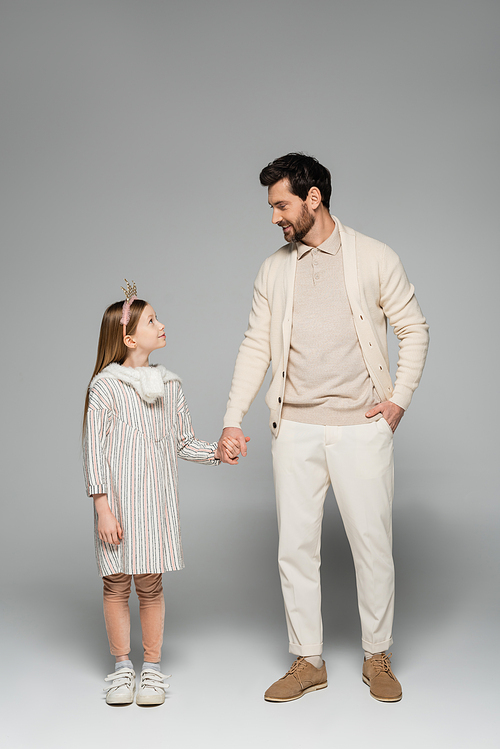 full length of happy man holding hands of kid in crown and standing with hand in pocket on grey