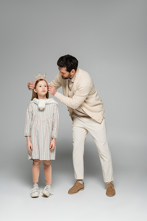 full length of bearded father adjusting crown of daughter in dress on grey