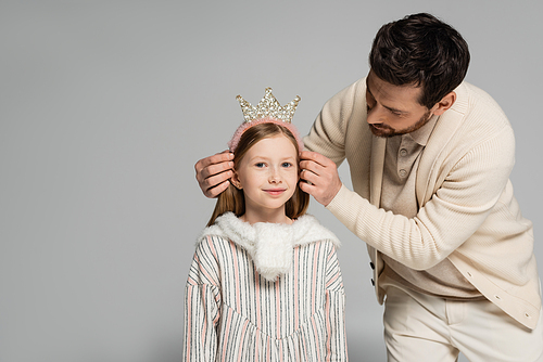 bearded father adjusting crown of smiling daughter in dress isolated on grey