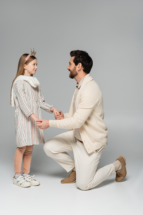 full length of happy man standing on knee and holding hands of daughter in dress and crown on grey