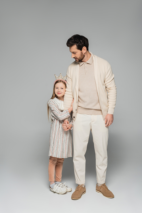 full length of happy girl in dress and crown hugging father in white outfit on grey