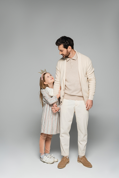 full length of cheerful girl in dress and crown hugging father in white outfit on grey