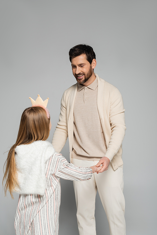 happy bearded man looking at daughter in crown while holding hands on grey