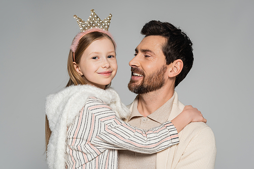 cheerful kid in crown looking at camera while hugging father isolated on grey