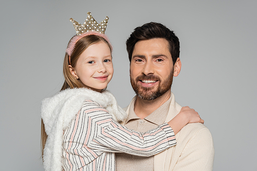 cheerful girl in crown looking at camera while hugging happy father isolated on grey