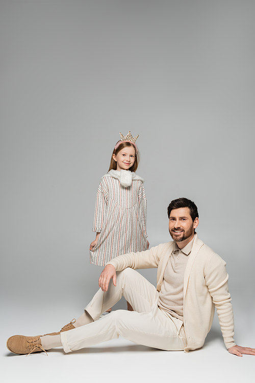 full length of cheerful girl in dress and crown standing near bearded father on grey