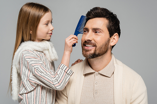 happy girl brushing hair of smiling and bearded father isolated on grey