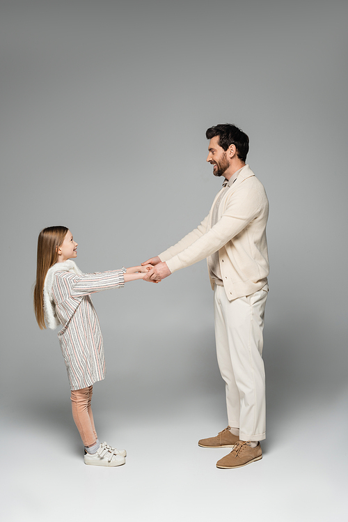 side view of happy father and daughter holding hands while looking at each other on grey