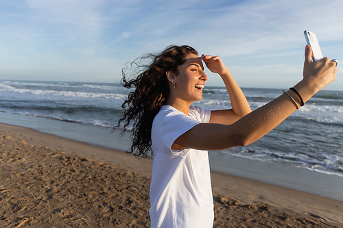 curly woman in white t-shirt taking selfie on smartphone on sandy beach in Barcelona