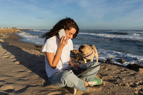 cheerful freelancer talking on smartphone while using laptop near pug dog on beach near sea in Spain