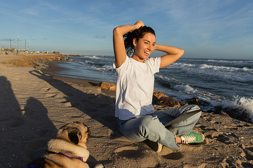 cheerful freelancer sitting with laptop and adjusting curly hair near pug dog on beach near sea in Spain